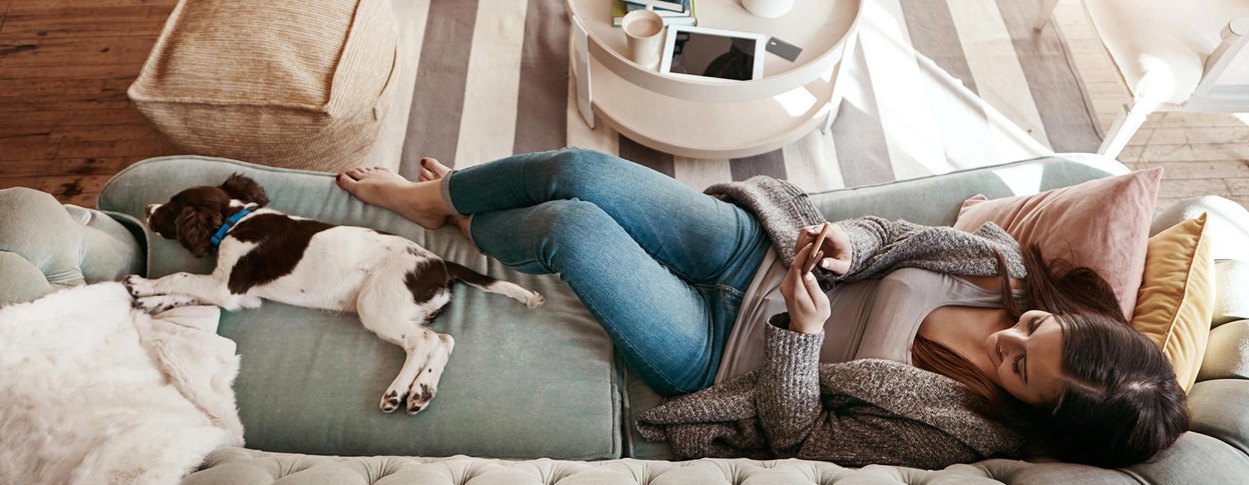 woman looks at phone while lying on a couch with her dog